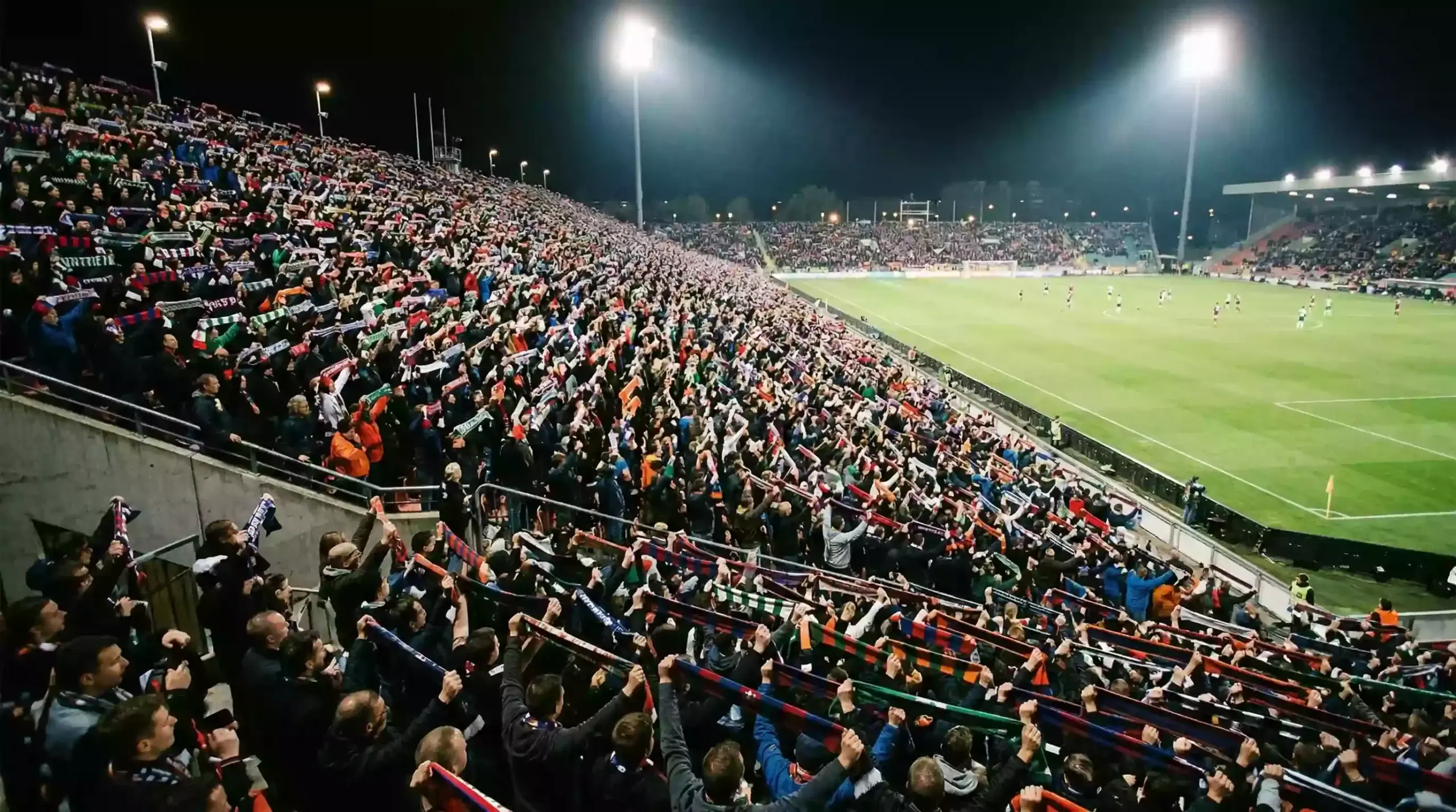 Grada llena de aficionados con bufandas en un estadio de fútbol durante un partido nocturno de Champions League